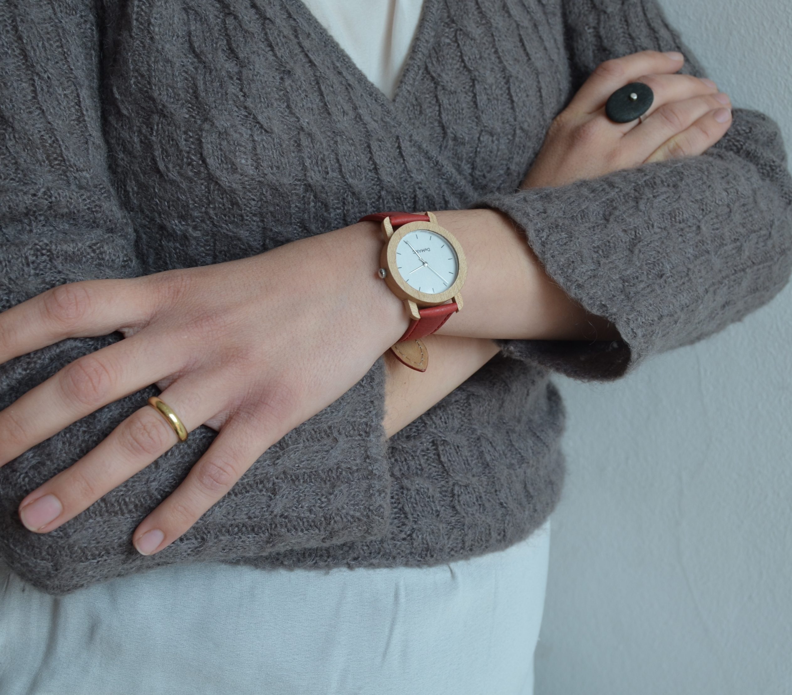 Detall d'una mà amb un rellotge de fusta i cuir vermell, amb un anell daurat, sobre una jaqueta de llana grisa.